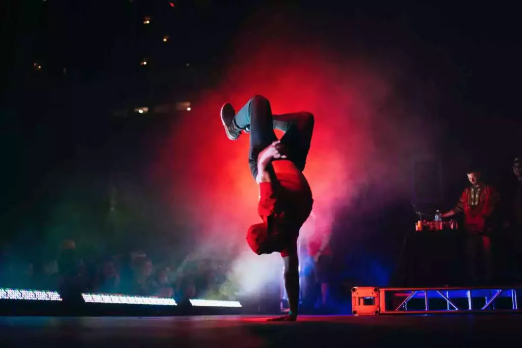 Man in a red hoodie break-dancing on a stage next to a music DJ.