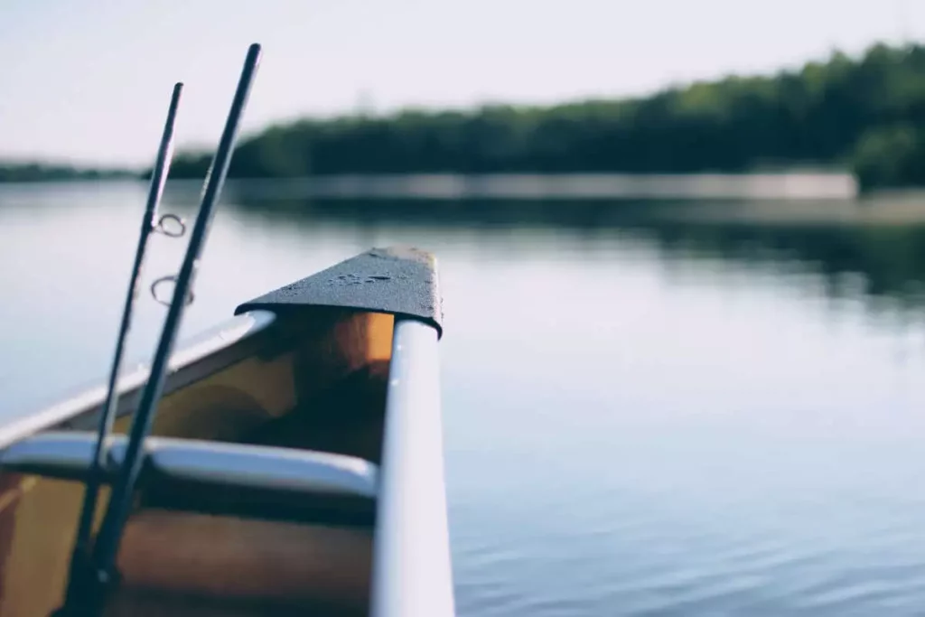 A pair of fishing poles resting on a small boat in a lake surrounded with green woods. 
