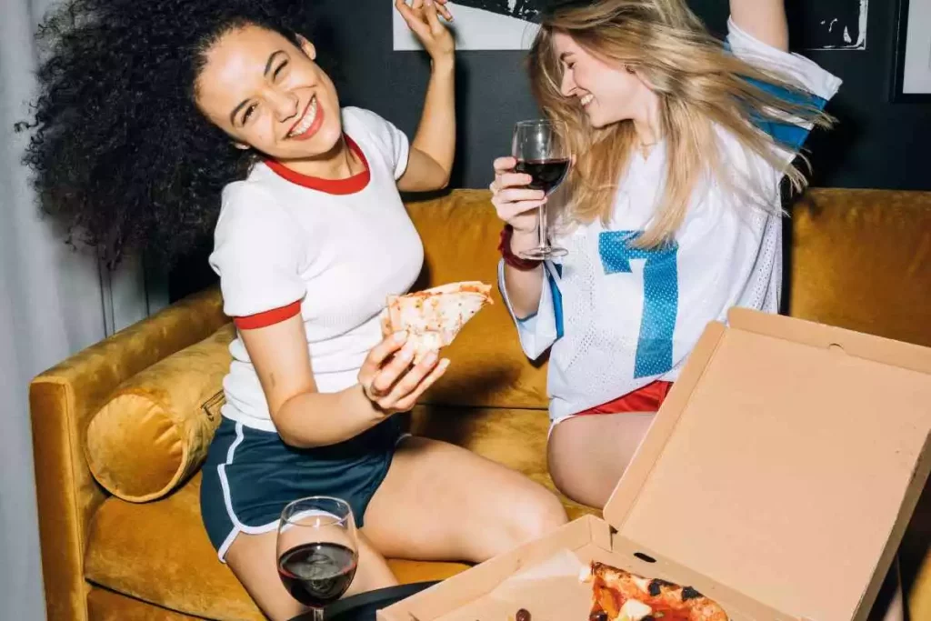 Two young girlfriends partying and having the time of their life while drinking wine and eating pizza during their girls night.