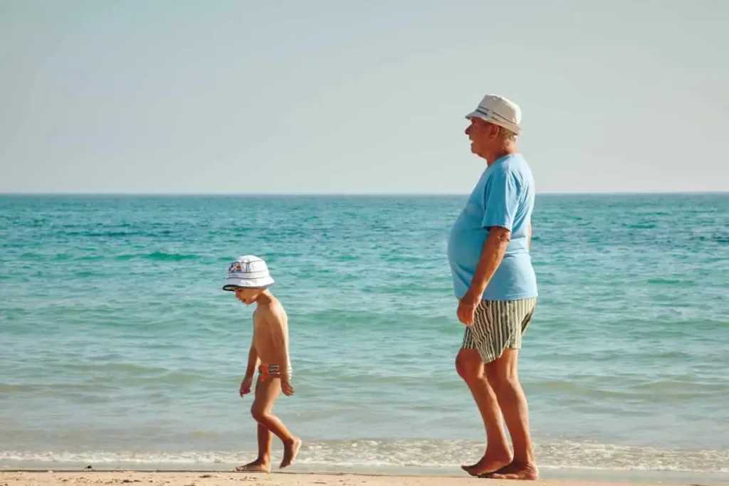 Grandfather walking with his young grandson on a sunny beach right next to the sea.