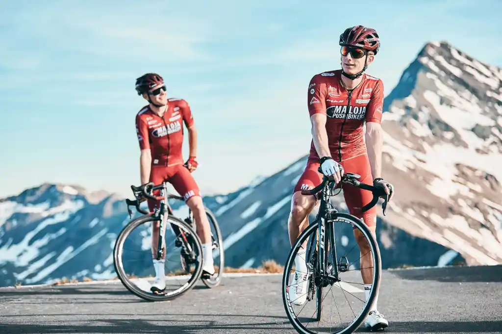 A photograph of two men cycling and a beautiful snowy mountain in the background. Cycling has many categories, however, mountain biking is the best one for short people.